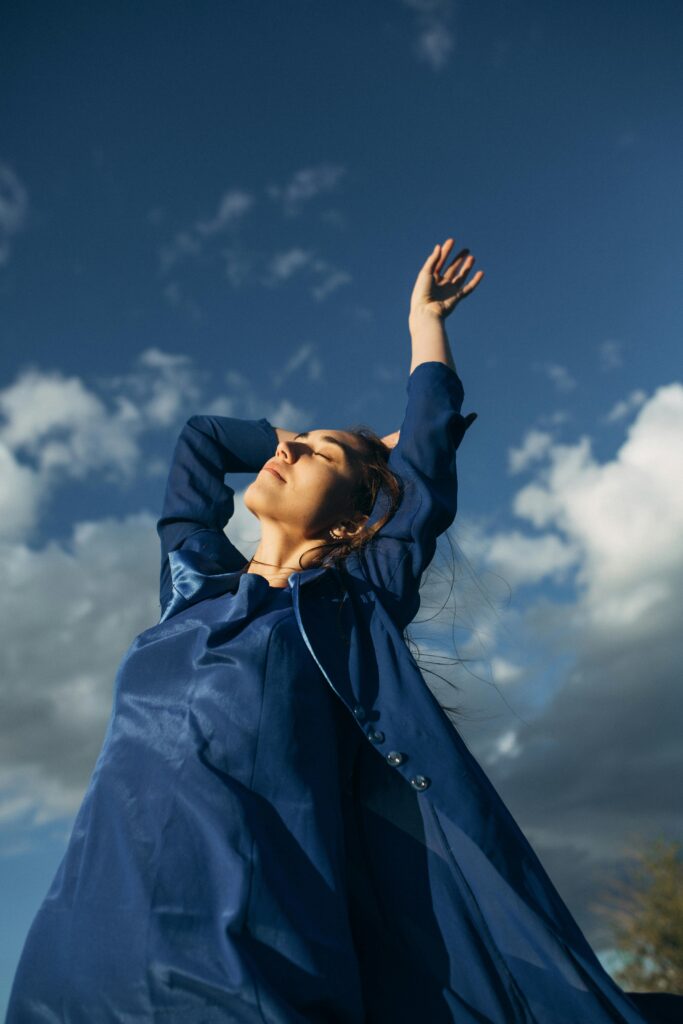 A woman in a blue dress enjoys the outdoors with arms raised under a clear sky.